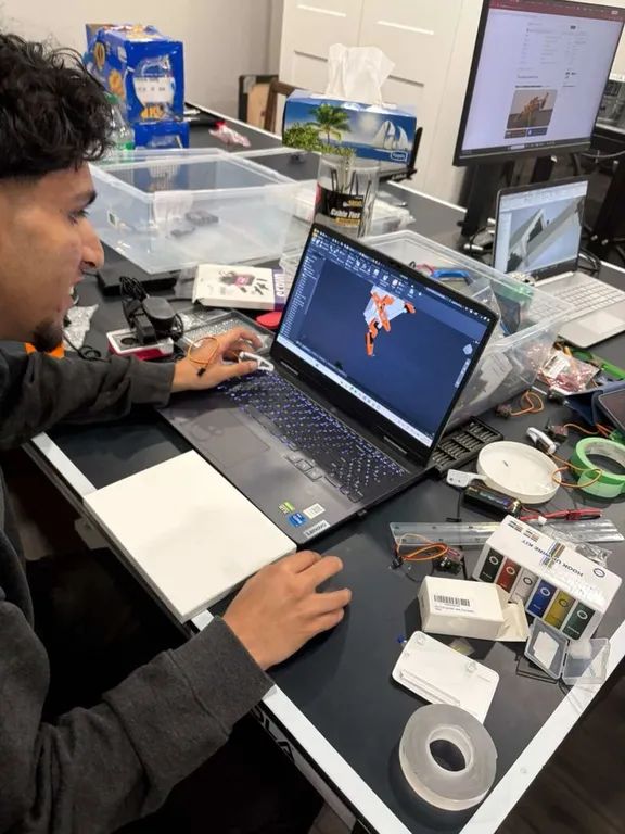 A laptop on a cluttered desk showing the quadruped assembly in Autodesk Inventor, body in white and legs in orange.