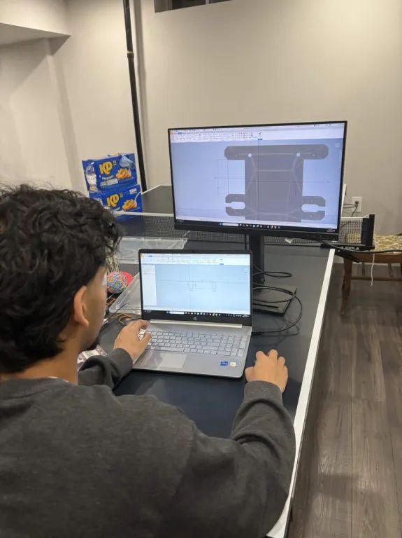 A monitor showing a 2D drawing of a chassis part with dimensions, a laptop below running the same CAD file.