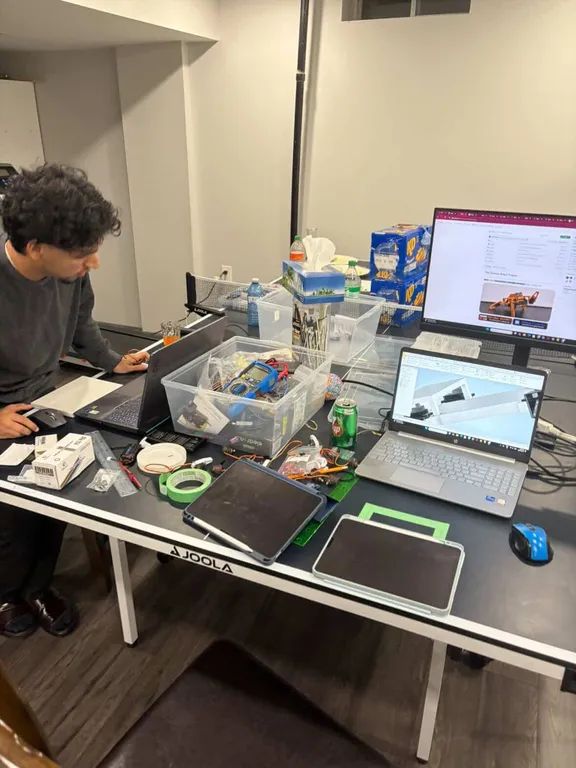 A wide view of the build space: a table top covered in components, tool boxes, two laptops and a monitor.