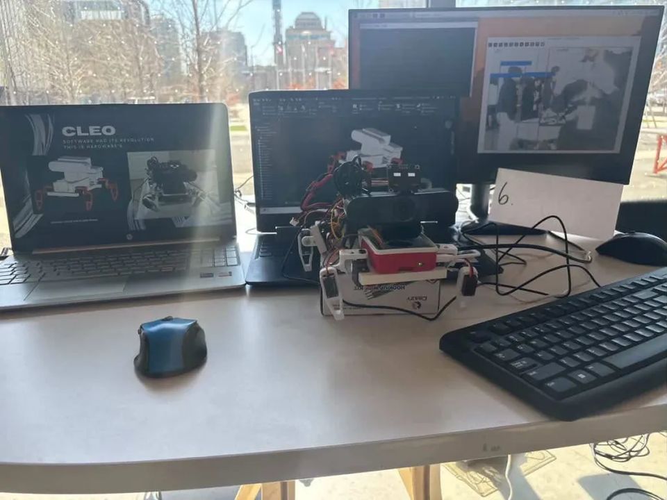 Demo table at the hackathon venue: a laptop showing the Cleo title slide, the robot in front of two screens, a numbered judging card beside it.