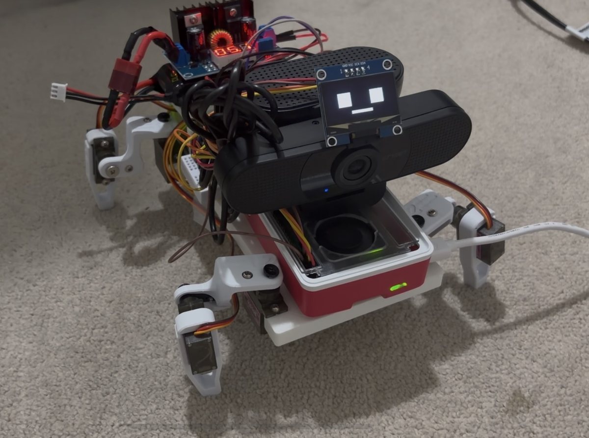 Cleo standing on a carpet: four white printed legs with servos at hip and knee, a Raspberry Pi in a red case, a webcam, and a small OLED showing a face.
