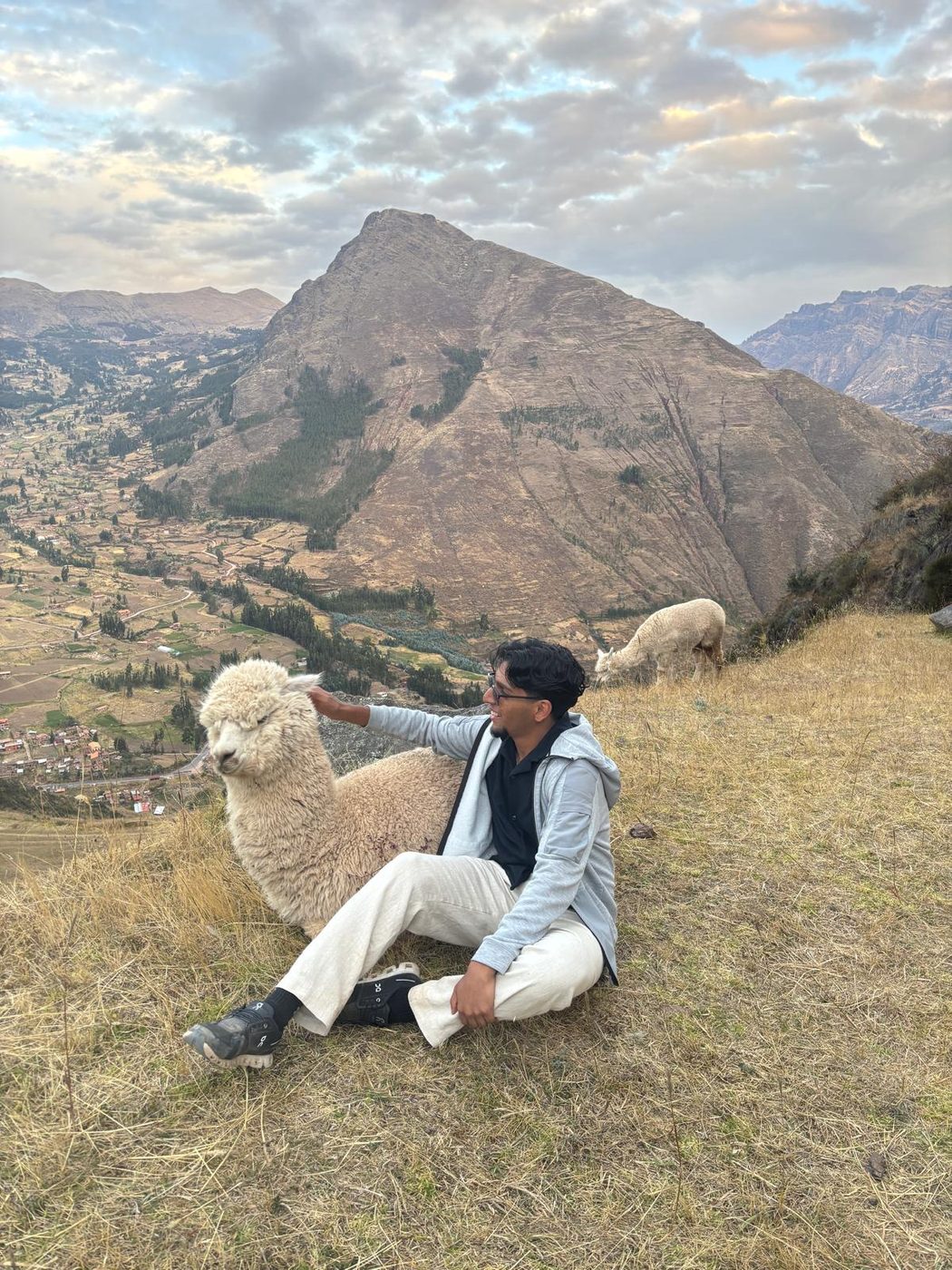 Sitting on a dry hillside above a wide valley with an alpaca standing over one shoulder.