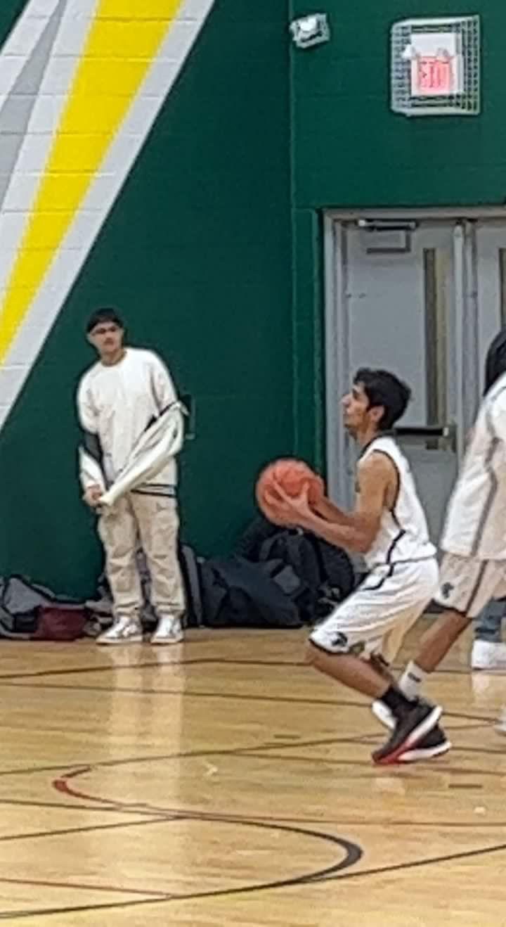 A basketball game in a school gym, a player in a white jersey driving along the baseline with the ball.