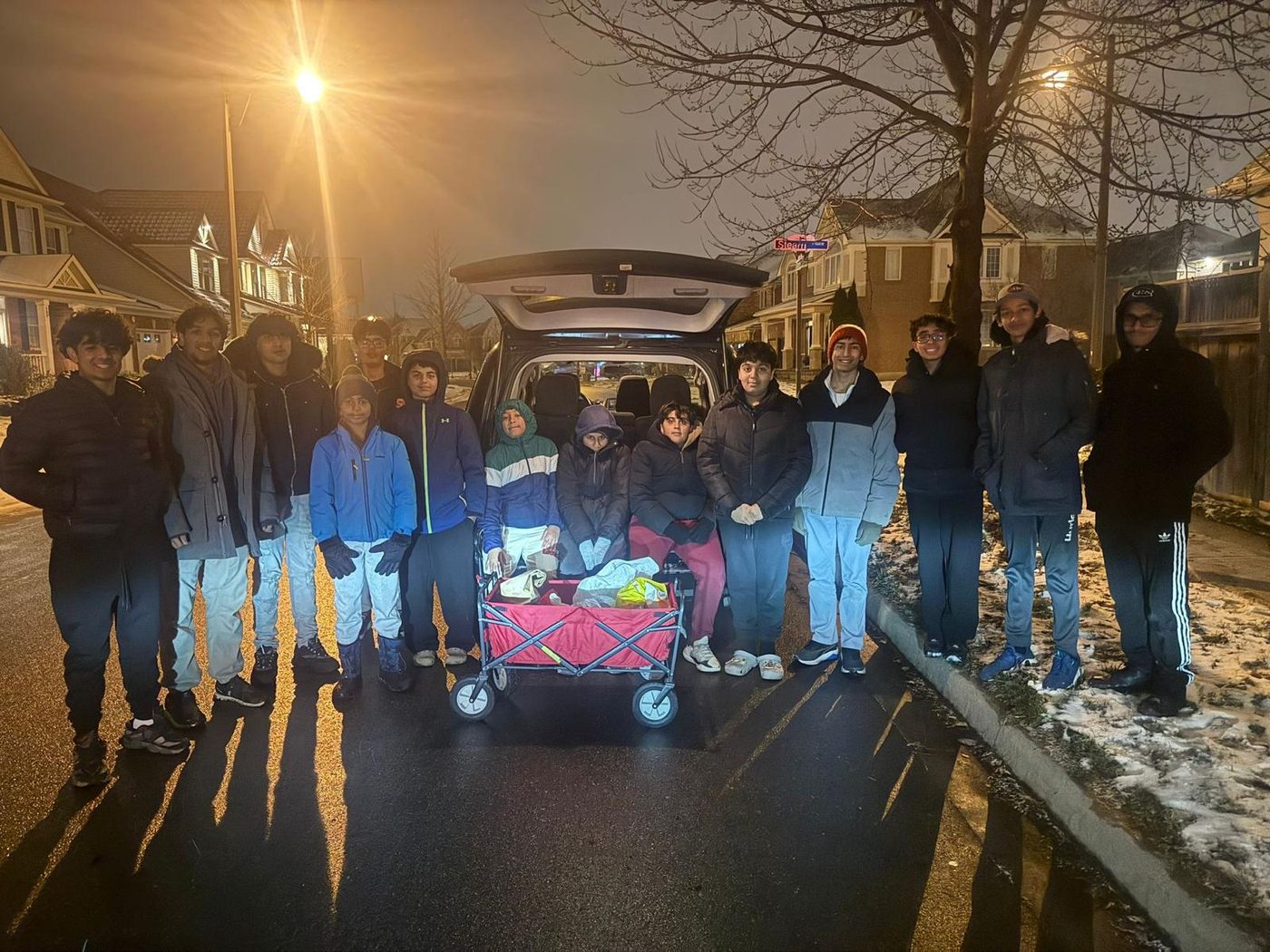 A group on a residential street at dusk behind an open hatchback and a loaded wagon, going door to door for donations.