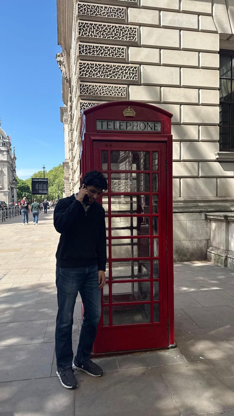 Leaning against a red telephone box on a London street in front of a stone building.
