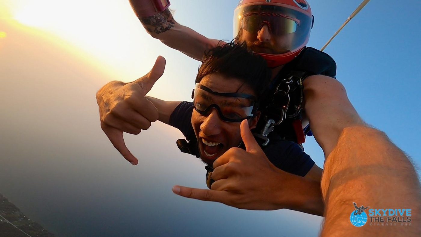 Tandem skydive in freefall, goggles on, throwing a shaka at the camera.