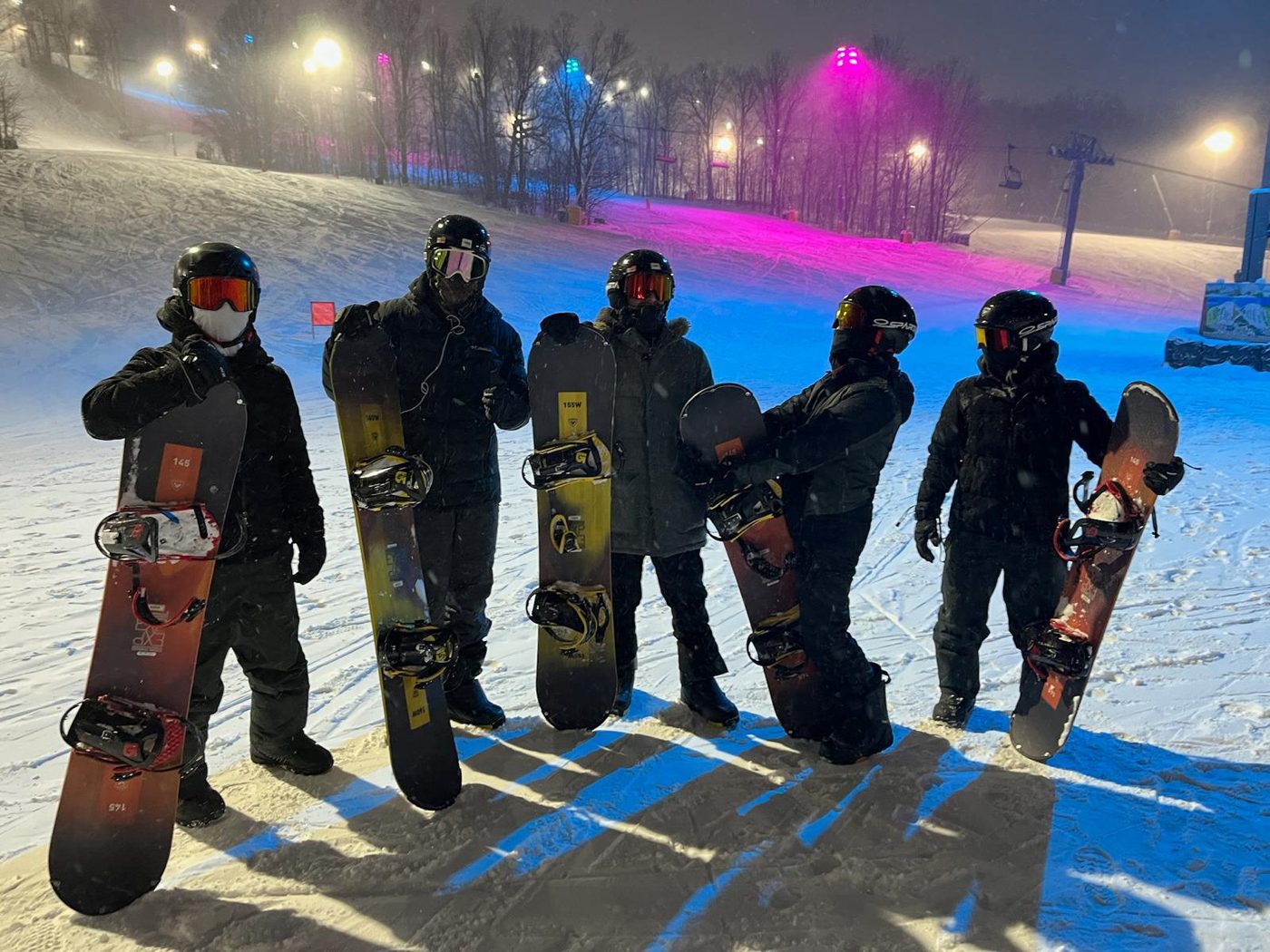 Five snowboarders lined up with their boards under the lights on a night run.