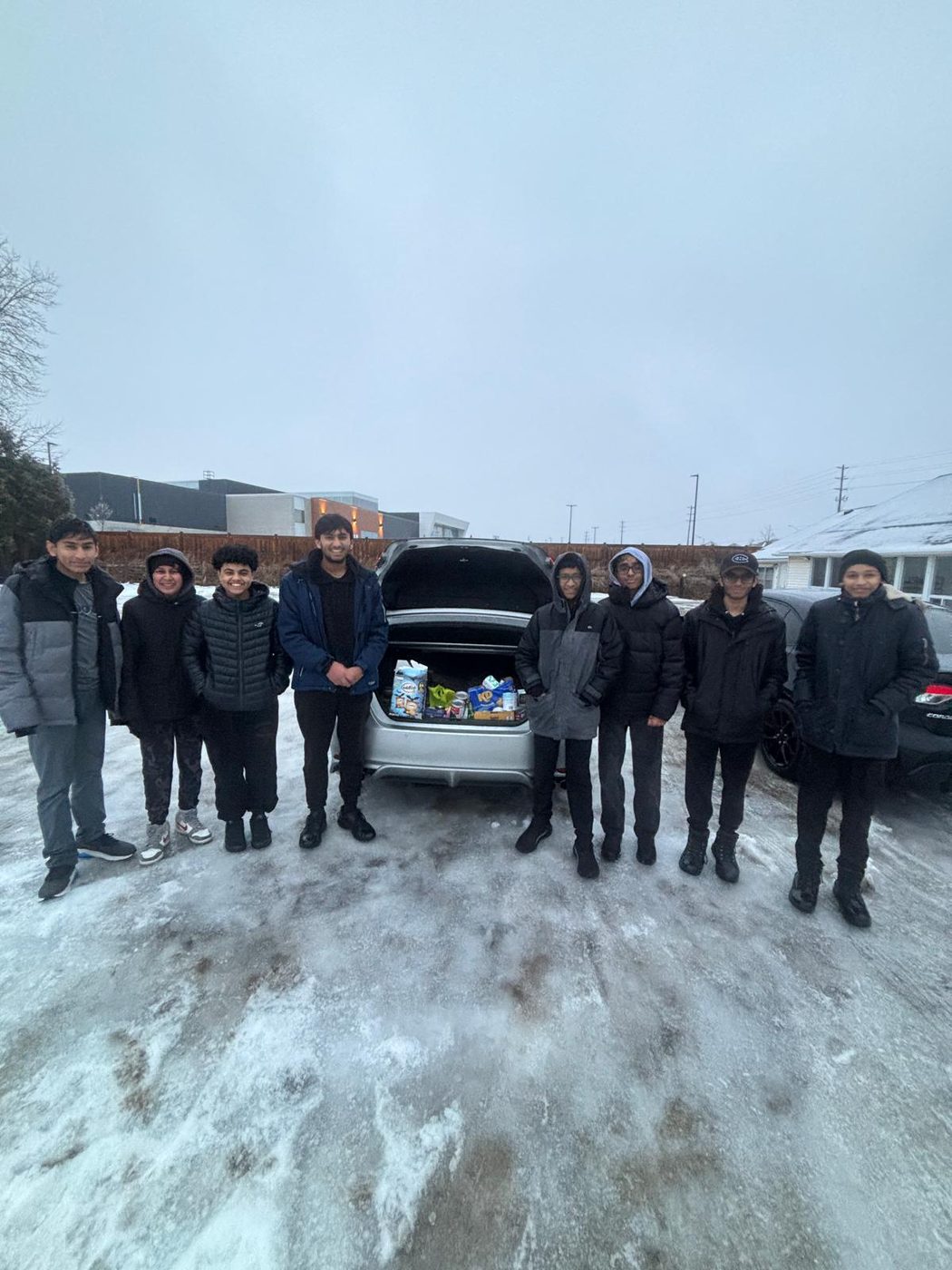 Nine of us standing in a snowy lot behind an open car boot packed with boxed and canned food.